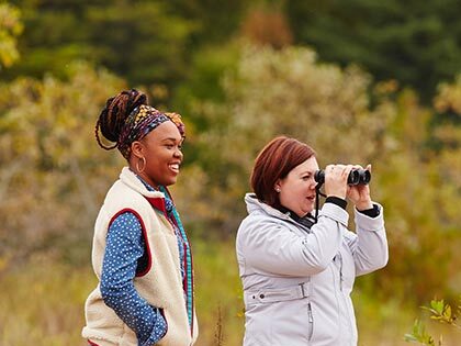 Two women standing in a grassy field as one looks through binoculars.