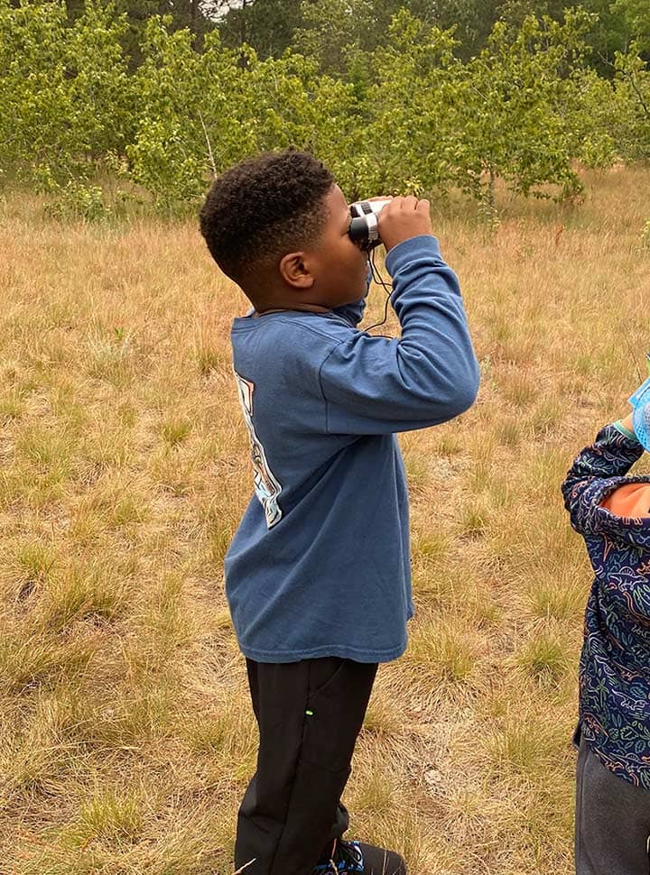 A boy in a field at summer camp looking through binoculars.