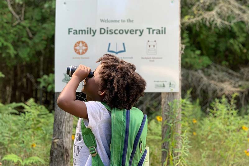 A child looks through binoculars with a sign that says Family Discover Trail behind her.